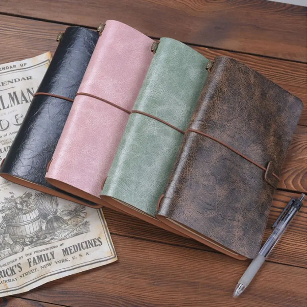 Four leather-bound journals in black, pink, green, and brown on a wooden surface with a pen and vintage newspaper.