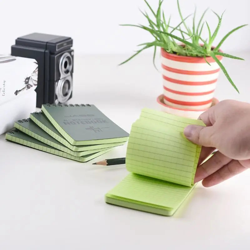 Hand holding a green notepad with stacked notebooks and a plant in the background