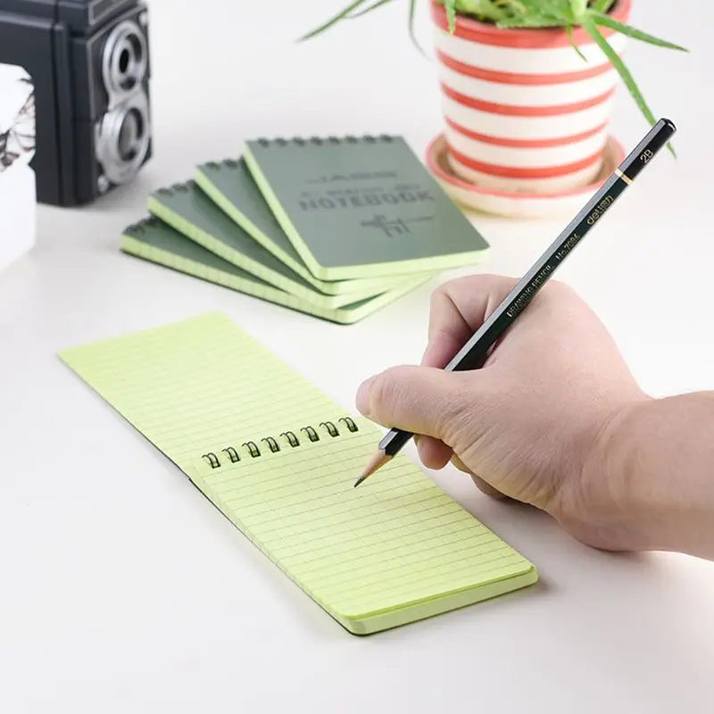 Hand holding a pencil over a green spiral-bound notepad with more notebooks and a plant in the background.