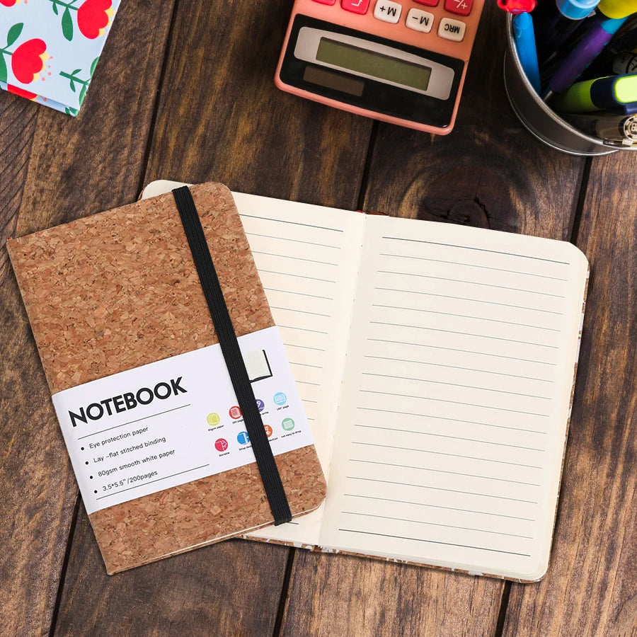 Open notebook with cork cover on a wooden surface with stationery items.