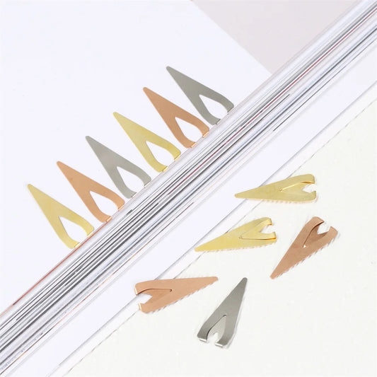 A set of metal bookmark clips in various colors including gold, silver, bronze, and copper, displayed on a white surface with a blurred background.