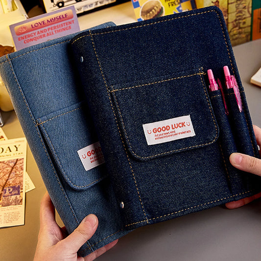 Blue denim notebook with 'Good Luck' label held by a person on a blurred background