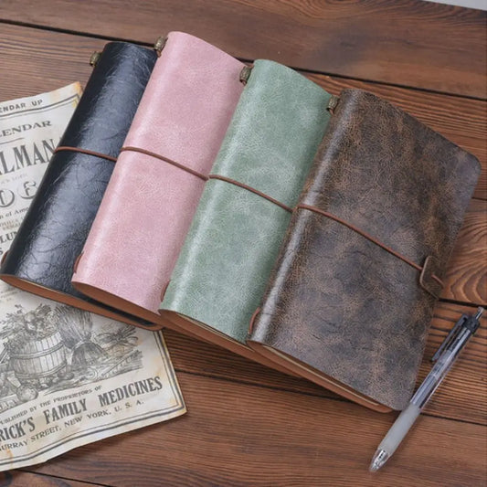 Four leather-bound journals in black, pink, green, and brown on a wooden surface with a pen and vintage newspaper.