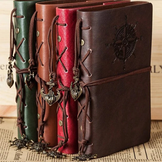 A collection of three retro-style pirate journal notebooks in brown, red, and green, each with a unique decorative strap and a metal medallion cover featuring an anchor design.