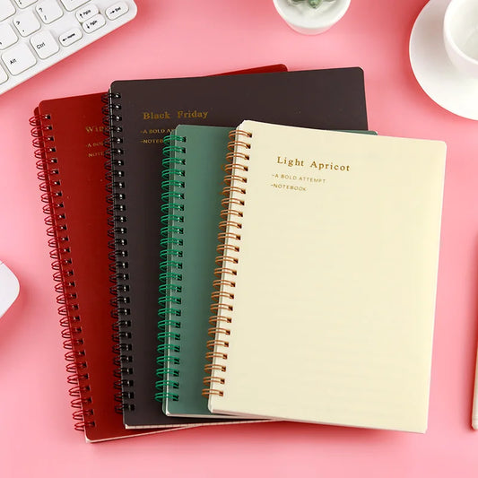 A collection of vintage spiral notebooks in black, red, green, and beige colors, displayed on a desk with a computer and other stationery items around.